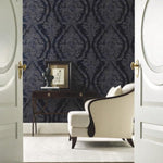 Double open doors reveal a stylish room adorned with Classic Charleston Damask Wallpaper in Blue (60 SqFt) by York Wallcoverings, a white upholstered armchair, and a wooden desk. On the desk sits a framed portrait and a small sculpture. The room features plush, light-colored carpeting.