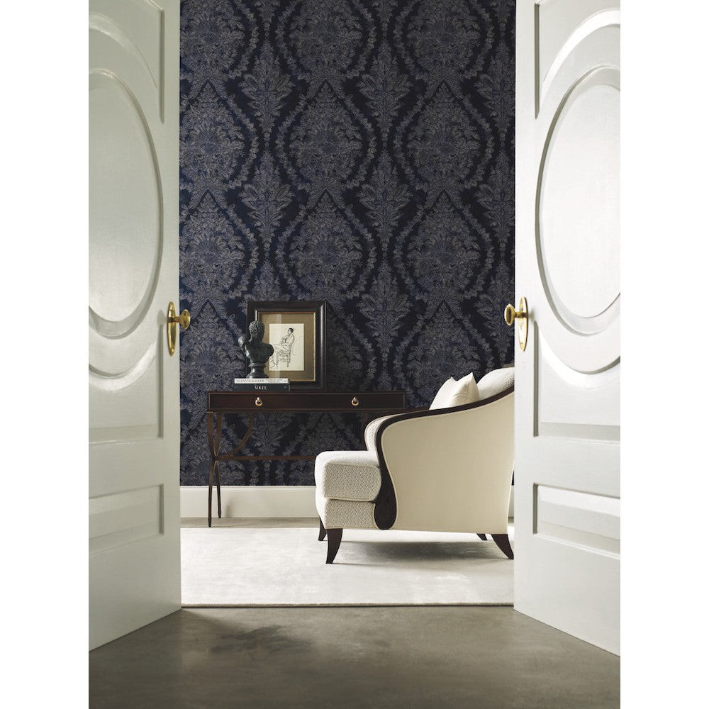 A luxurious room is seen through double doors featuring York Wallcoverings' Grey Charleston Damask Elegant Wallpaper and a white armchair. A wooden console table displays artwork and a sculpture, with light-colored carpet flooring.