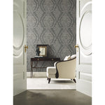 An elegant room with York Wallcoverings Classic Charleston Damask Wallpaper in Blue (60 SqFt), viewed through open double doors. It features a cream-colored armchair, a dark wooden side table with drawers, and decorative items, including a framed picture and a small statue. A light rug covers part of the floor.