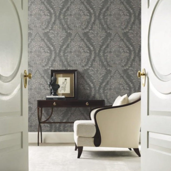 Through open white double doors, a room features an elegant, white armchair alongside a dark brown console table. The walls are adorned with York Wallcoverings' Grey Charleston Damask Elegant Wallpaper, adding sophistication. The table displays a framed picture and small bust for added grace.