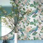 A decorative, washable wallpaper with peacock and floral patterns adorns a wall. A round mirror reflects part of the room, and a white vase containing branches with green leaves and white flowers sits on a blue ledge below the mirror. The overall decor, enhanced by the colorful plumage, is light and airy thanks to the Garden Rose and Plume (60 SqFt) wallpaper from York Wallcoverings.