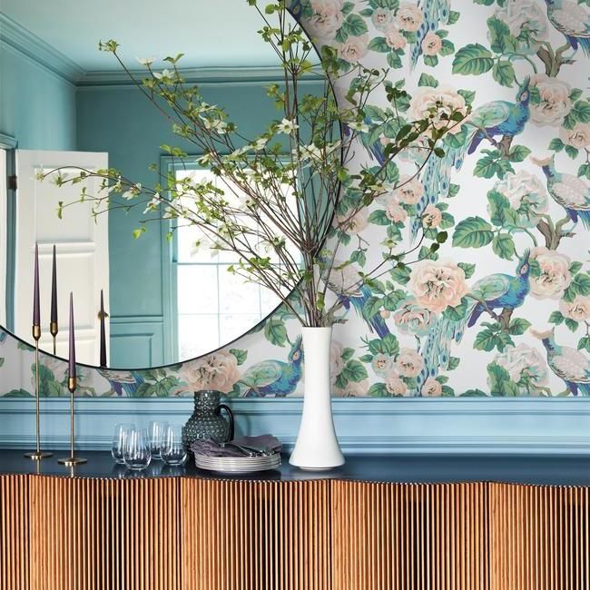The decorative room features York Wallcoverings' Garden Plume Wallpaper (60 SqFt) with colorful plumage and peacock designs, a round mirror, and a sideboard with vertical wood paneling. On the sideboard are candles, glasses, plates, and a tall white vase holding branches with green leaves. The room has soft blue accents.