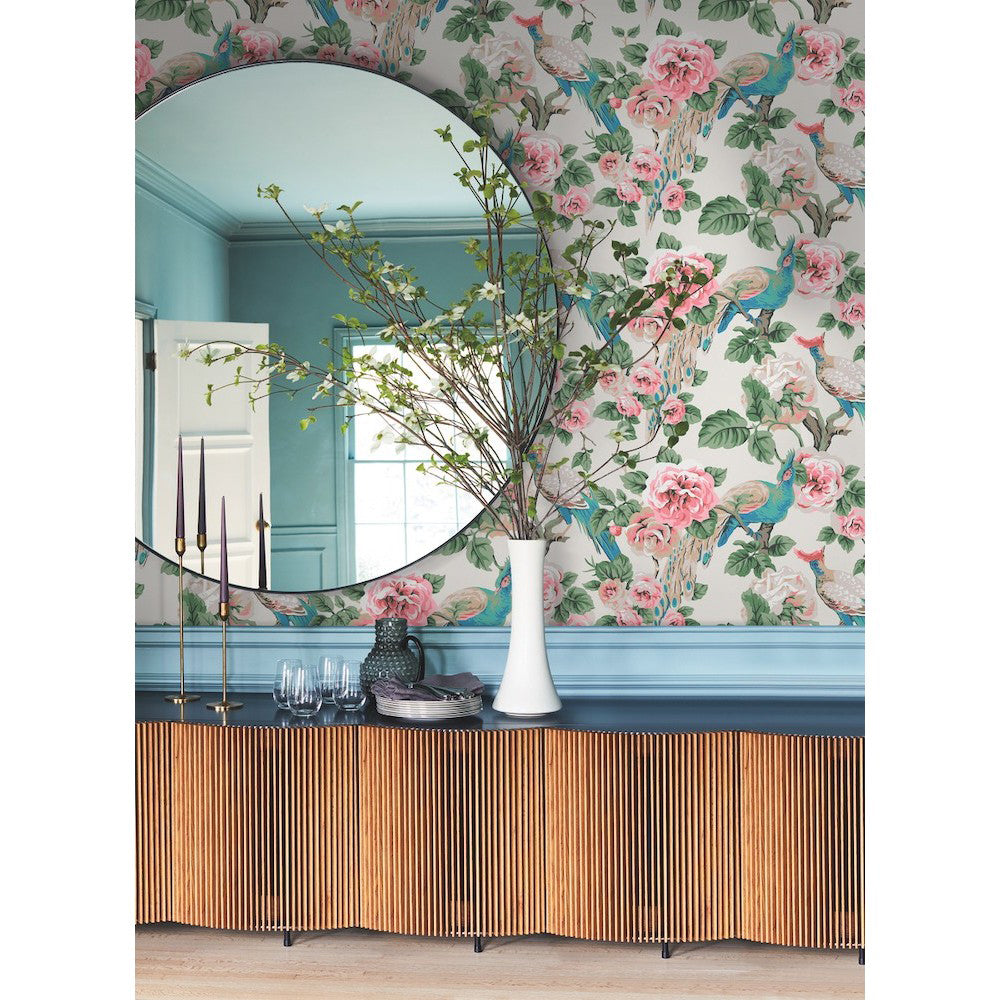 A decorative sideboard with vertical wooden slats sits beneath a large round mirror. The wall is adorned in York Wallcoverings' Garden Rose and Plume (60 SqFt) featuring pink roses and blue accents in a large scale pattern. A tall white vase with branches stands on the sideboard, alongside candles and glassware.