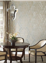 A cozy dining area with a dark round table, four cream-colored cushioned chairs, and a metallic chandelier above. A potted orchid with branches sits on the table. York Wallcoverings Classic Charleston Damask Wallpaper in Blue (60 SqFt) and a window with tan curtains complete the elegant setting.