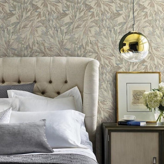 A cozy bedroom features a beige tufted headboard, a bed with white and gray pillows and bedding, a side table with a framed picture, a vase of white hydrangeas, and a book. A modern spherical pendant light hangs above, with the wall adorned in Rainforest Leaves (56 SqFt) wallpaper by York Wallcoverings.