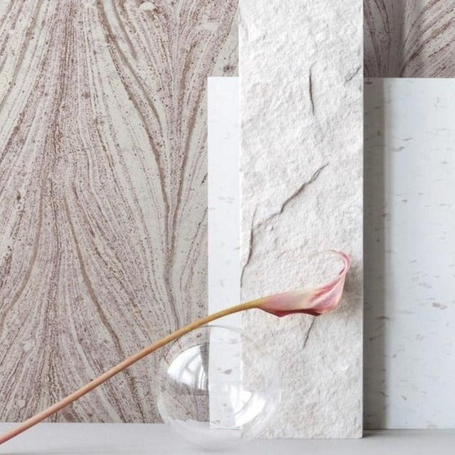 A minimalist still life image featuring a delicate pink calla lily arching out of a small, clear glass vase. The backdrop consists of three textured, vertically arranged white panels with varying patterns, set against an Ebru Marble Texture (56 SqFt) surface from York Wallcoverings with swirling designs.