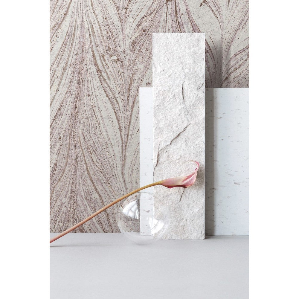 A minimalist composition featuring a single pink calla lily bending over a small, clear, rounded vase. Behind the vase are three vertically arranged textured panels in varying shades of off-white, set against a subtly patterned beige **Ebru Marble (56 SqFt) by York Wallcoverings** background.