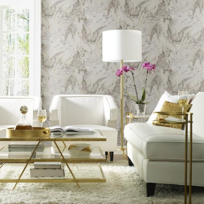 Modern living room with luxury wallcoverings featuring Oil & Marble (56 SqFt) by York Wallcoverings, white furniture, and gold accents. A glass coffee table holds a decanter and books, while a pink orchid sits on a side table with a white lamp. Sunlight streams through a large window, highlighting the cozy ambiance.